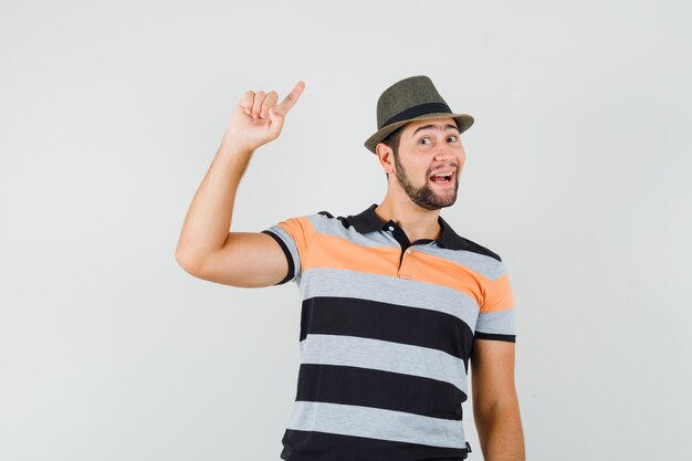 Jonge man in t-shirt, hoed wijst naar de zijkant met vinger omhoog en kijkt nieuwsgierig, vooraanzicht.