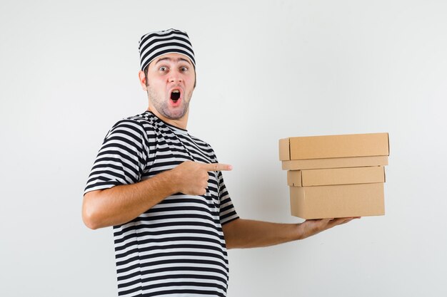 Jonge man in t-shirt, hoed wijzend op kartonnen dozen en kijkt verbaasd, vooraanzicht.