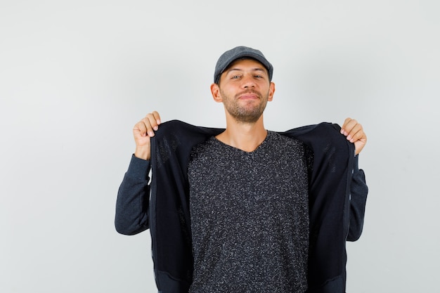 Jonge man in t-shirt, jas, hoed die zijn jas bevestigt en zelfverzekerd kijkt, vooraanzicht.