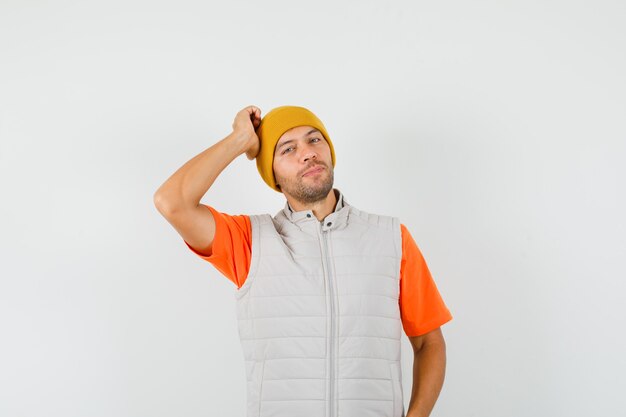 Jonge man in t-shirt, jas, hoed poseren met de hand op het hoofd en ziet er schattig uit