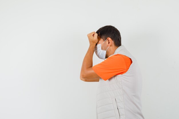 Jonge man in t-shirt, jas, masker vuisten drukken op voorhoofd en op zoek gespannen.