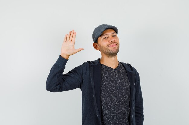 Jonge man in t-shirt, jas, pet zwaaiende hand voor begroeting en glimlachen, vooraanzicht.