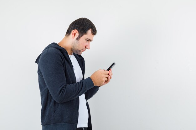 Jonge man in t-shirt, jasje met behulp van mobiele telefoon en op zoek naar druk, vooraanzicht.