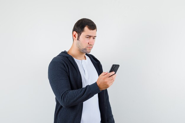 Jonge man in t-shirt, jasje met behulp van mobiele telefoon en op zoek naar druk, vooraanzicht.