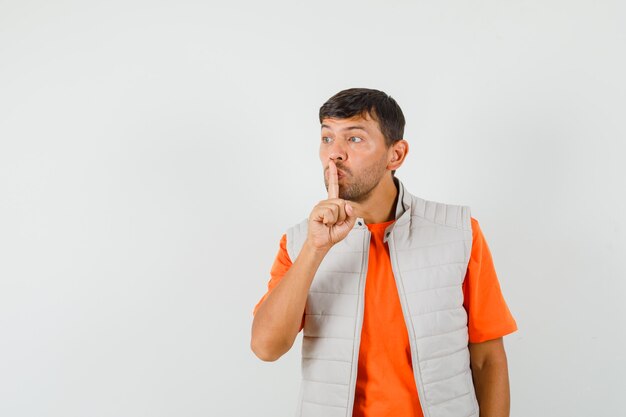 Jonge man in t-shirt, jasje met stilte gebaar en op zoek voorzichtig, vooraanzicht.