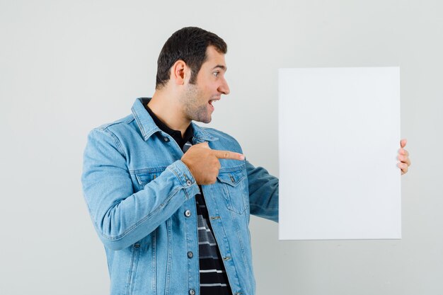 Jonge man in t-shirt, jasje wijzend op leeg canvas en op zoek vrolijk