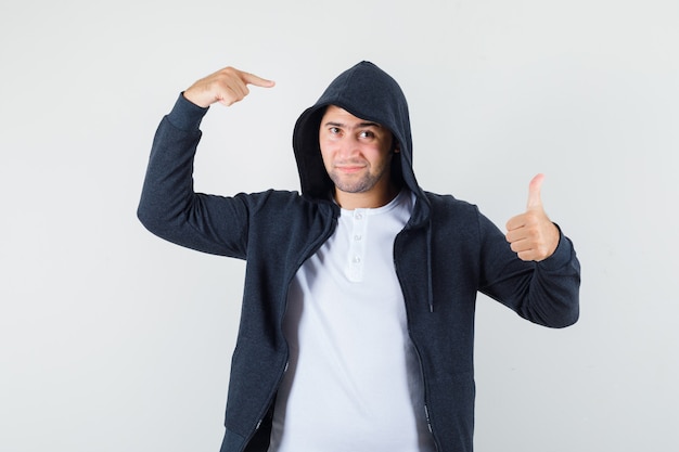 Gratis foto jonge man in t-shirt, jasje wijzend op zijn kap, duim opdagen en op zoek naar zelfverzekerd, vooraanzicht.