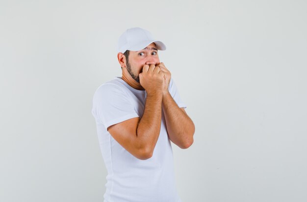 Jonge man in t-shirt, pet die de mond bedekt met zijn vuisten en doodsbang kijkt.