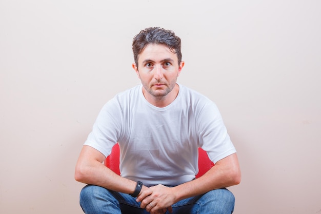 Jonge man in t-shirt, spijkerbroek zittend op een stoel en verbaasd kijkend