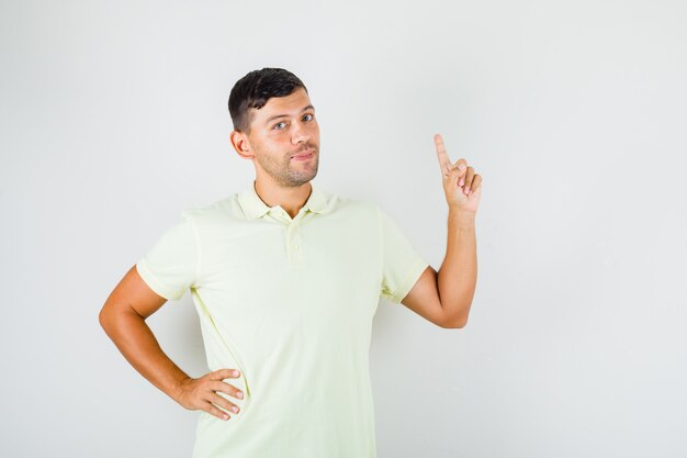 Jonge man in t-shirt wijsvinger met hand op de taille verhogen en er zelfverzekerd uitzien