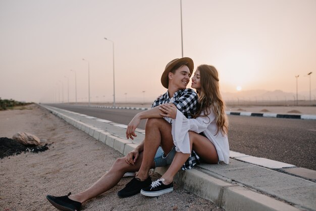 Jonge man in trendy hoed kijken met liefde naar zijn sierlijke vriendin in wit overhemd, terwijl rust na wandeling. Aantal reizigers die dichtbij de weg zitten en zachtjes omhelzen met zonsondergang