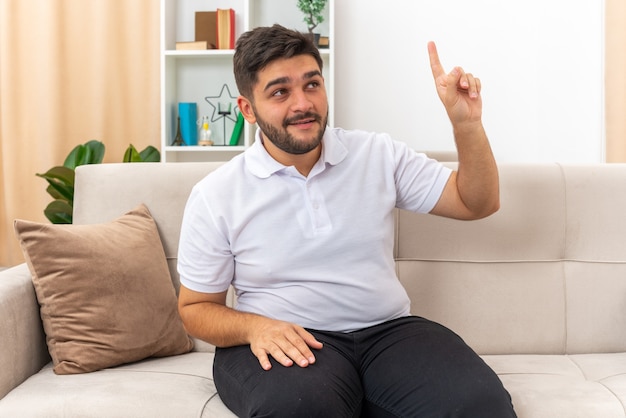 Jonge man in vrijetijdskleding die blij en verrast opkijkt en wijsvinger laat zien met een nieuw goed idee zittend op een bank in een lichte woonkamer