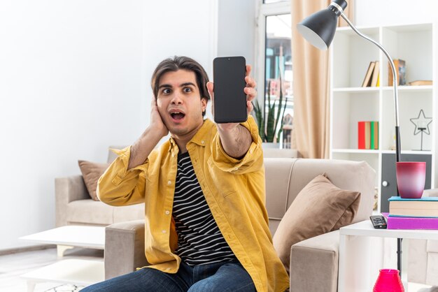Jonge man in vrijetijdskleding die een smartphone laat zien die verbaasd en verrast op de stoel zit in een lichte woonkamer