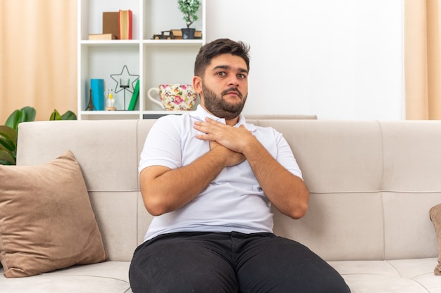 Jonge man in vrijetijdskleding die er bezorgd en verward uitziet met handen op zijn borst zittend op een bank in lichte woonkamer