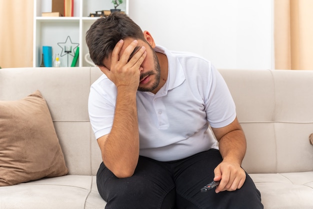 Gratis foto jonge man in vrijetijdskleding die er depressief uitziet en ogen bedekt met de hand zittend op een bank in een lichte woonkamer