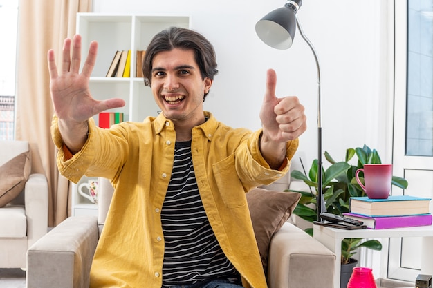 Jonge man in vrijetijdskleding die er gelukkig en vrolijk uitziet en nummer zes toont met vingers op de stoel in een lichte woonkamer