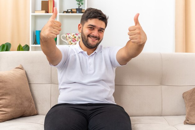 Jonge man in vrijetijdskleding die er gelukkig uitziet en vrolijk lacht, met zijn duimen omhoog zittend op een bank in een lichte woonkamer