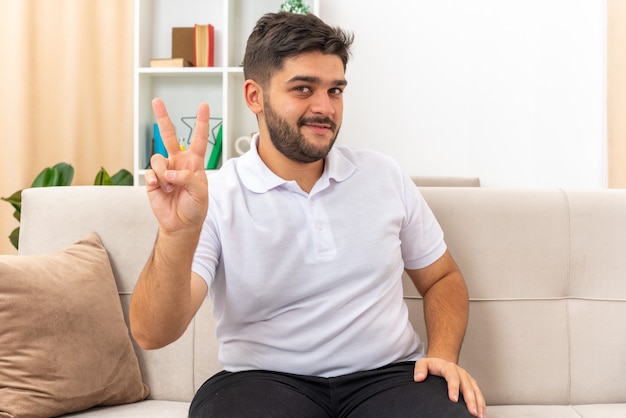 Jonge man in vrijetijdskleding die lacht en vrolijk een v-teken toont terwijl hij op een bank zit in een lichte woonkamer