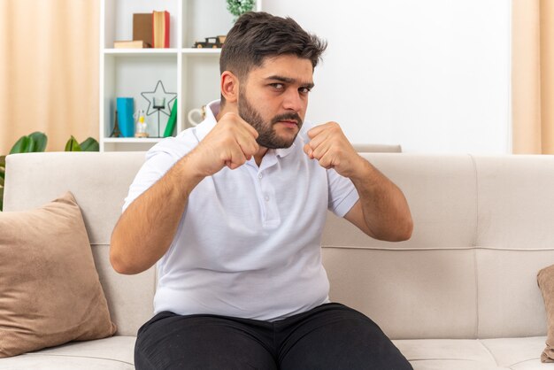 Jonge man in vrijetijdskleding die met gebalde vuisten kijkt als een winnaar die op een bank in een lichte woonkamer zit