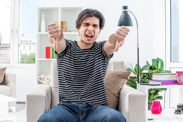 Jonge man in vrijetijdskleding die opgewonden en ontevreden schreeuwt en duimen naar beneden laat zien terwijl hij op de stoel zit in een lichte woonkamer