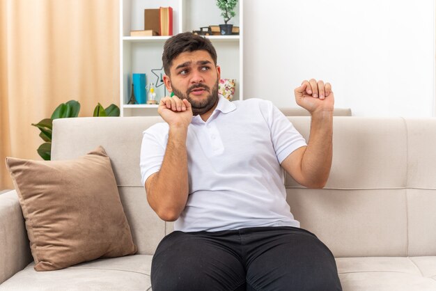 Jonge man in vrijetijdskleding die opzij kijkt, bezorgd om een verdedigingsgebaar te maken zittend op een bank in een lichte woonkamer