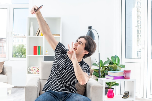 Jonge man in vrijetijdskleding die selfie maakt met smartphone, blij en positief met v-teken zittend op de stoel in lichte woonkamer