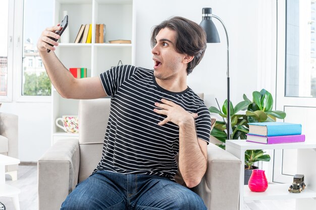 Jonge man in vrijetijdskleding die selfie maakt met smartphone en er verbaasd en verrast uitziet terwijl hij op de stoel zit in een lichte woonkamer