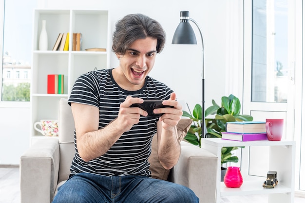 Jonge man in vrijetijdskleding die spelletjes speelt met smartphone, blij en opgewonden zittend op de stoel in een lichte woonkamer