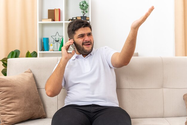 Jonge man in vrijetijdskleding die verward en ontevreden kijkt terwijl hij op een mobiele telefoon praat en zijn arm in verontwaardiging opheft, zittend op een bank in een lichte woonkamer