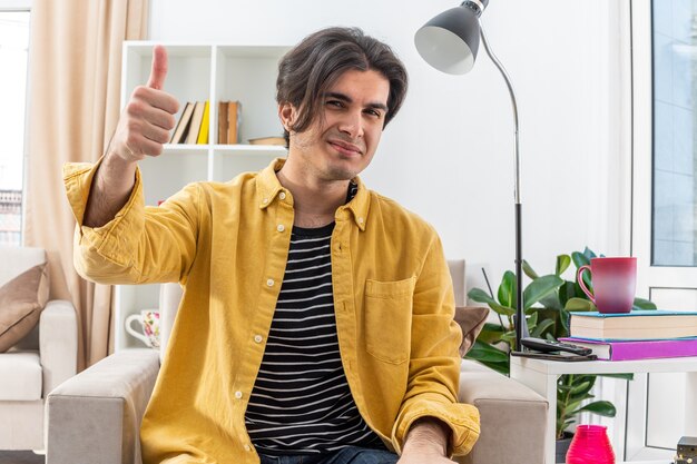 Jonge man in vrijetijdskleding die vrolijk lacht en duimen laat zien terwijl hij op de stoel zit in een lichte woonkamer