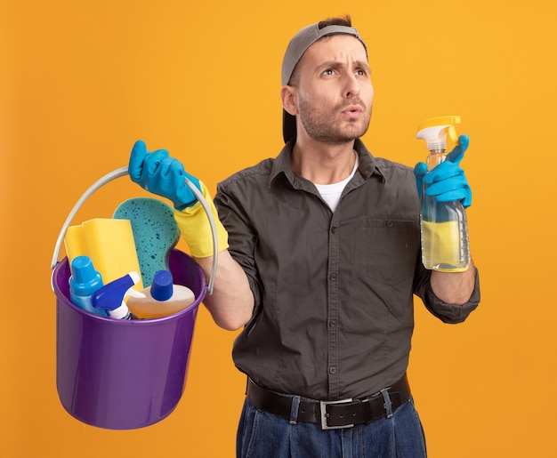 Jonge man in vrijetijdskleding en pet met emmer met schoonmaakgereedschap opzij kijken verbaasd staande over oranje muur