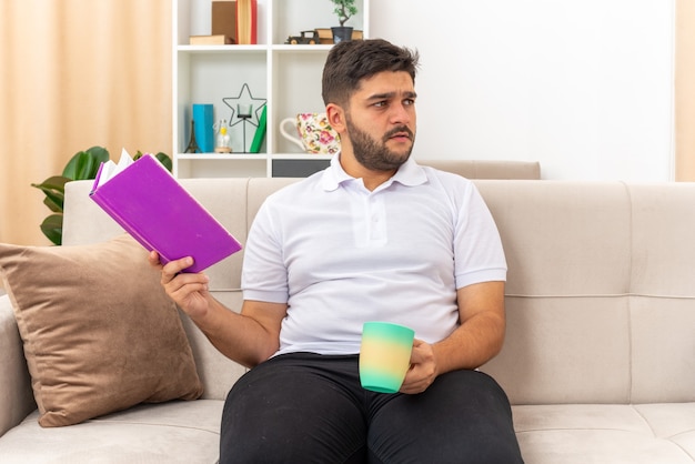 Jonge man in vrijetijdskleding met boek en beker opzij kijkend verbaasd zittend op een bank in lichte woonkamer