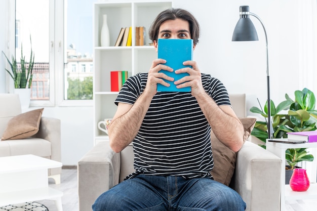 Jonge man in vrijetijdskleding met boek voor zijn gezicht, blij en positief zittend op de stoel in lichte woonkamer