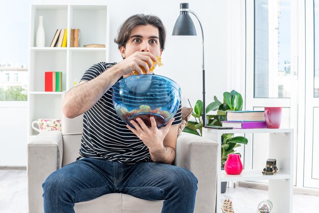 Jonge man in vrijetijdskleding met een kom chips etend en verbaasd en verrast zittend op de stoel in een lichte woonkamer living