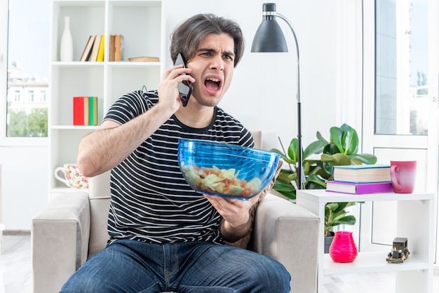 Jonge man in vrijetijdskleding met een kom chips schreeuwend ontevreden terwijl hij op een mobiele telefoon praat terwijl hij op de stoel zit in een lichte woonkamer