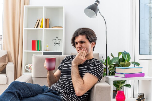 Jonge man in vrijetijdskleding met een kop hete thee die er onwel uitziet en zijn wang aanraakt met kiespijn zittend op de stoel in een lichte woonkamer