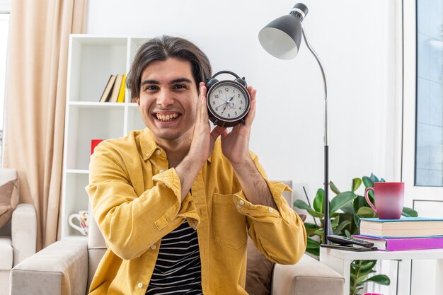 Jonge man in vrijetijdskleding met een wekker die blij en vrolijk glimlacht, breeduit zittend op de stoel in een lichte woonkamer