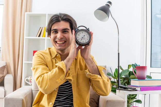 Jonge man in vrijetijdskleding met een wekker die blij en vrolijk glimlacht, breeduit zittend op de stoel in een lichte woonkamer