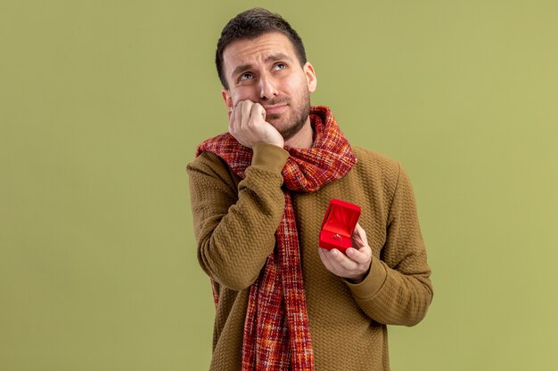 jonge man in vrijetijdskleding met sjaal om nek met rode doos met verlovingsring opzoeken bezorgd Valentijnsdag concept staande over groene muur