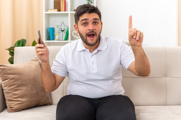 Jonge man in vrijetijdskleding met smartphone die er blij en verrast uitziet en laat zien dat wijsvinger een nieuw idee heeft zittend op een bank in een lichte woonkamer light