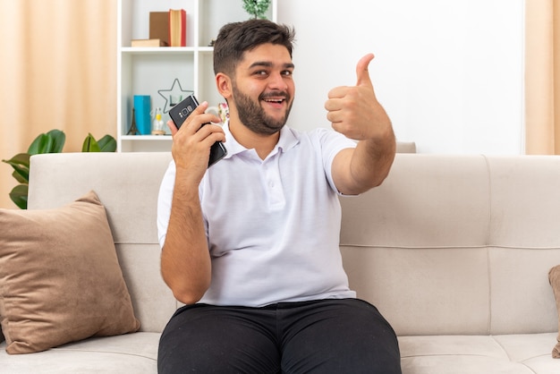 Jonge man in vrijetijdskleding met smartphone die er gelukkig en vrolijk uitziet en duimen opsteekt glimlachend breed zittend op een bank in lichte woonkamer living