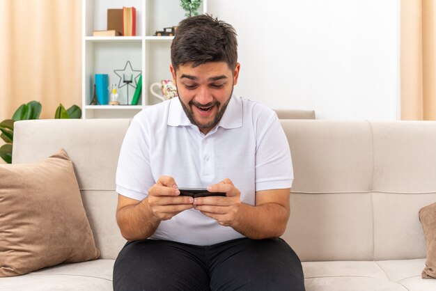 Jonge man in vrijetijdskleding met smartphone die spelletjes speelt, blij en vrolijk zittend op een bank in een lichte woonkamer