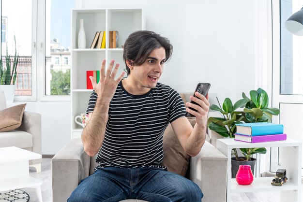 Jonge man in vrijetijdskleding met smartphone die verbaasd en verrast op de stoel zit in een lichte woonkamer