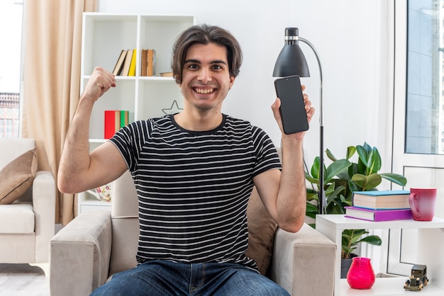 Jonge man in vrijetijdskleding met smartphone die vuist opsteekt als een winnaar, blij en positief zittend op de stoel in een lichte woonkamer