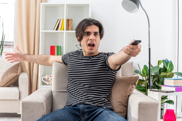 Jonge man in vrijetijdskleding met tv op afstand blij en verrast kijkend naar zijn favoriete film zittend op de stoel in lichte woonkamer