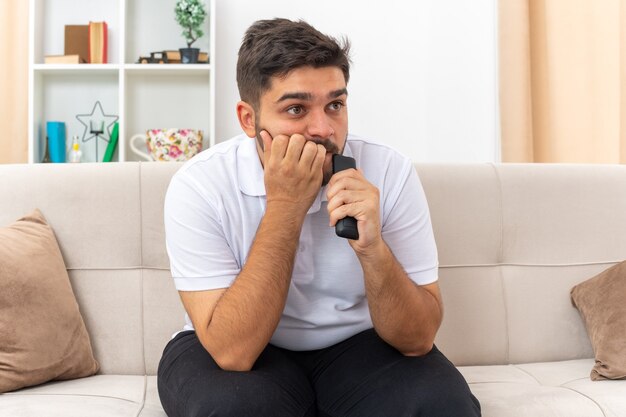 Jonge man in vrijetijdskleding met tv op afstand tv kijkend er gestrest en nerveus uitziend weekend thuis zittend op een bank in lichte woonkamer