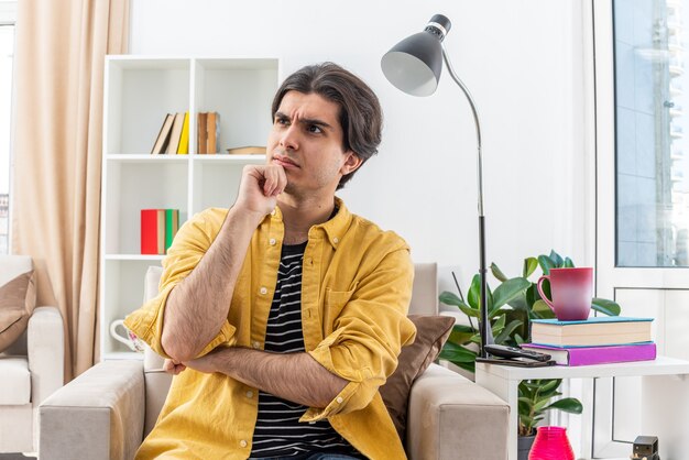 Jonge man in vrijetijdskleding opzij kijkend met peinzende uitdrukking met de hand op de kin denkend zittend op de stoel in een lichte woonkamer light