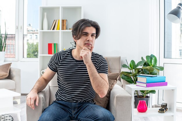 Jonge man in vrijetijdskleding zittend op de stoel met peinzende uitdrukking met hand op kin in lichte woonkamer