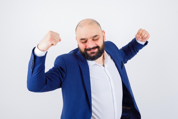 Jonge man in wit overhemd, jas die winnaargebaar toont en er gelukkig uitziet, vooraanzicht.