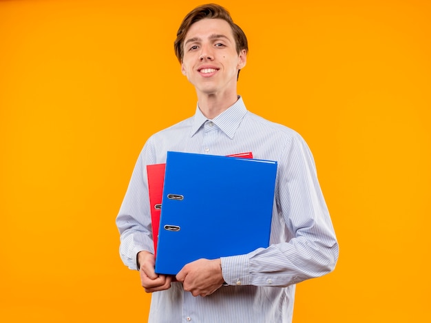 Jonge man in wit overhemd met mappen kijken met glimlach op gezicht blij en zelfverzekerd staande over oranje muur